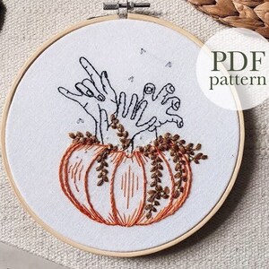 May include: A PDF pattern for an embroidery design featuring a pumpkin with hands reaching out of the top. The pumpkin is orange with brown stems and leaves. The hands are black and white. The text "PDF pattern" is written in black on a white background.