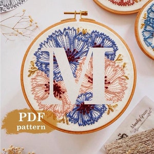 Può includere: Ricamo floreale su cerchio con la lettera "M" bianca, circondata da fiori blu e pesca. Il testo "PDF pattern" è visibile. Altri cerchi con lettere e disegni floreali sono sullo sfondo.