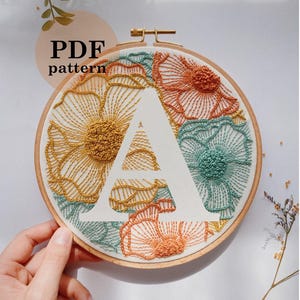 May include: Embroidered floral hoop art featuring the letter "A" in white. The design includes flowers in shades of yellow, orange, and teal. The text "PDF pattern" is visible. The text "mockup of stitched design with the letter" is also visible.