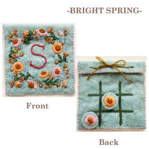 May include: A light blue felt pouch with a floral embroidery design and the letter "S" on the front. The back of the pouch has a noughts and crosses board with two embroidered circles.