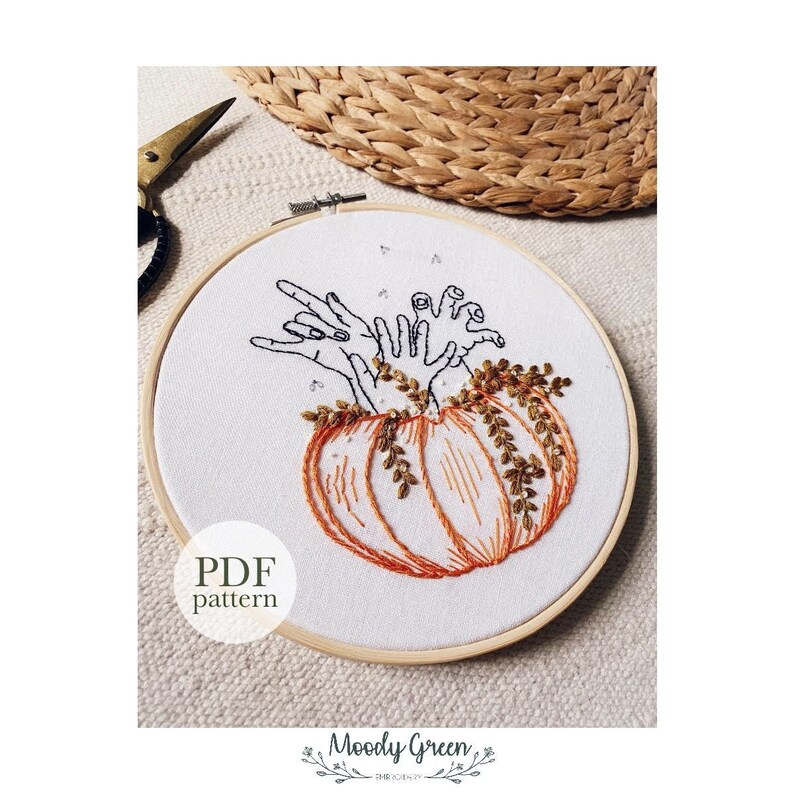 May include: Embroidered hoop art featuring a pumpkin with multiple hands reaching out from the top. The pumpkin is orange with brown stems and leaves. The hands are black and white. The background is white. The text "PDF pattern" is below the hoop.