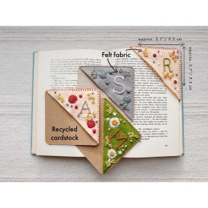 May include: Four felt fabric corner bookmarks with embroidered letters "S", "A", "M", and "R" in different colors. The bookmarks are 3.7 inches (9.5 cm) wide and are placed on a book with the text "Recycled cardstock" visible.