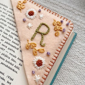 May include: A pink felt bookmark with a green embroidered letter "R" surrounded by white flowers, yellow leaves, and purple beads.