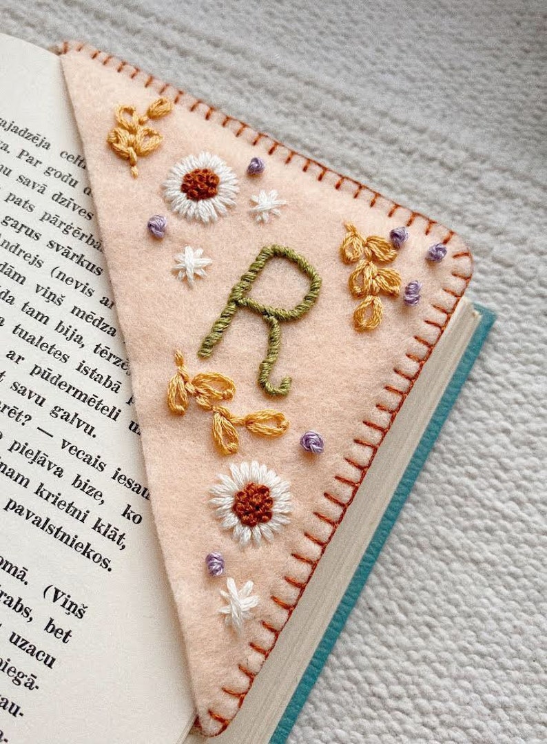 May include: A pink felt bookmark with a green embroidered letter "R" surrounded by white flowers, yellow leaves, and purple beads.