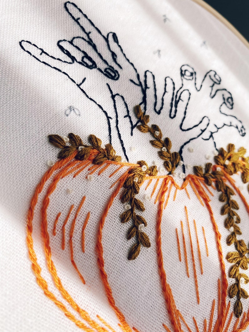 May include: Embroidered design of two hands reaching towards a pair of pumpkins with green leaves. The pumpkins are orange and the hands are black.