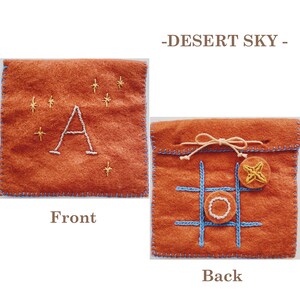 May include: A felt pouch with a brown background and blue stitching. The front of the pouch has a white letter A embroidered on it with yellow stars. The back of the pouch has a tic-tac-toe board embroidered on it with a yellow star and a white circle.