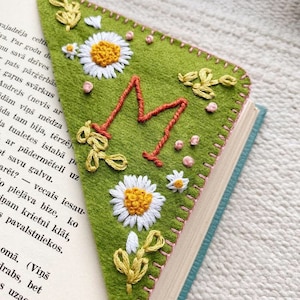 May include: A green felt bookmark with a large embroidered letter "M" in orange and pink thread. The bookmark is decorated with white daisies and green leaves, all embroidered in colorful thread.