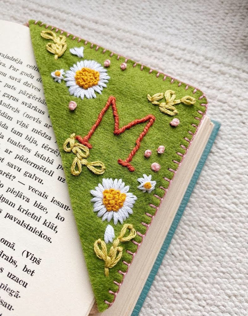 May include: A green felt bookmark with a large embroidered letter "M" in orange and pink thread. The bookmark is decorated with white daisies and green leaves, all embroidered in colorful thread.