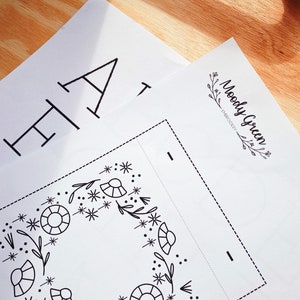 May include: A black and white embroidery pattern featuring a floral wreath design. The pattern is on a white sheet of paper with the text "Moody Green Embroidery" in the top right corner.