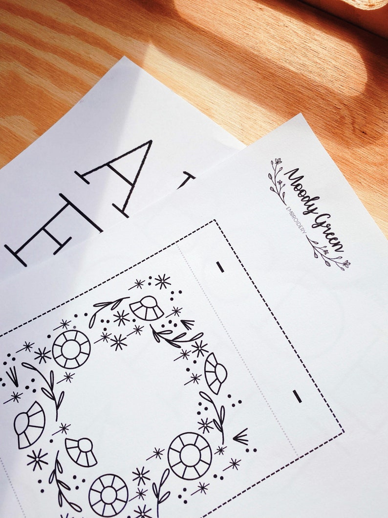 May include: A black and white embroidery pattern featuring a floral wreath design. The pattern is on a white sheet of paper with the text "Moody Green Embroidery" in the top right corner.
