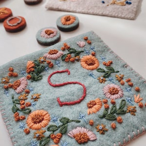 May include: A light blue felt pouch with a pink stitched border. The pouch is embroidered with a floral design in shades of orange, pink, green, and blue. The letter "S" is embroidered in red in the centre of the pouch.