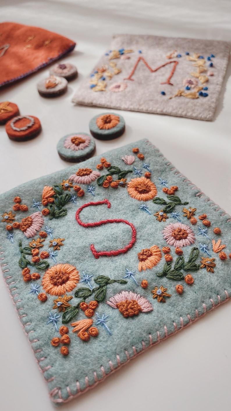 May include: A light blue felt pouch with a pink stitched border. The pouch is embroidered with a floral design in shades of orange, pink, green, and blue. The letter "S" is embroidered in red in the centre of the pouch.
