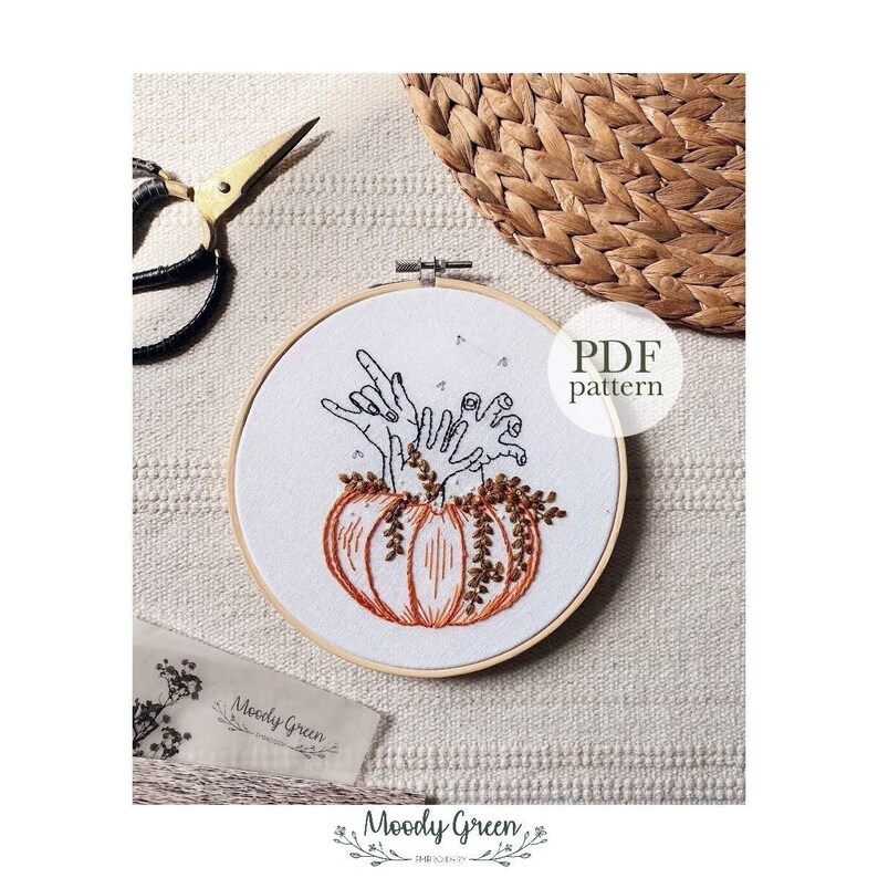 May include: A PDF pattern for an embroidery design featuring a pumpkin with hands reaching out of the top. The pumpkin is orange with brown stems and leaves. The hands are black and white. The text "PDF pattern" is written in black on a white background.