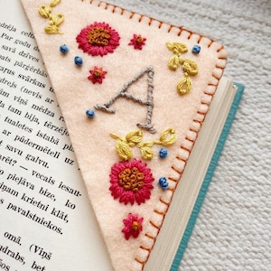 May include: A pink felt bookmark with a hand-stitched letter A, flowers, and leaves. The bookmark is placed on a book with blue pages.