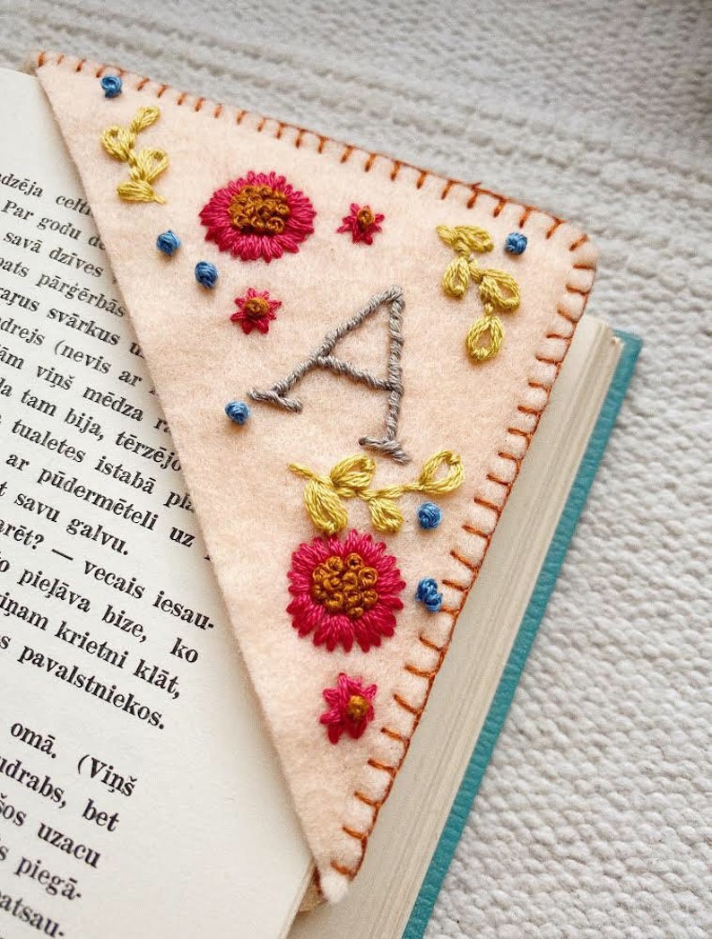 May include: A pink felt bookmark with a hand-stitched letter A, flowers, and leaves. The bookmark is placed on a book with blue pages.