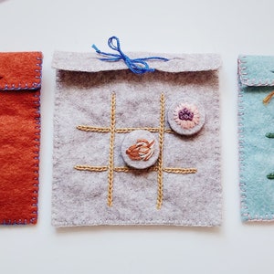 May include: Three felt tic-tac-toe game pouches with different coloured felt and embroidery. The pouches are tied with twine and have buttons for game pieces.