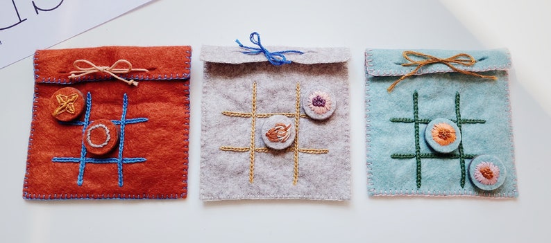 May include: Three felt tic-tac-toe game pouches with different coloured felt and embroidery. The pouches are tied with twine and have buttons for game pieces.