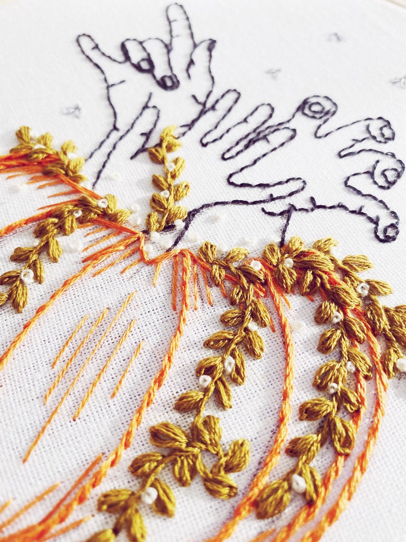 May include: Embroidered pumpkin with green leaves and white beads, with two hands in the air making the 'I love you' sign language gesture.