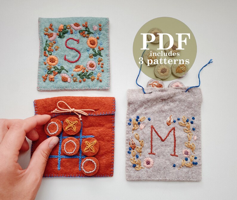 May include: Three felt pouches with embroidery. The first pouch is light blue with a floral wreath and the letter "S" in the centre. The second pouch is orange with a noughts and crosses board and nine felt circles. The third pouch is grey with a floral wreath and the letter "M" in the centre. The text "PDF includes 3 patterns" is in a circle above the pouches.