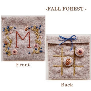 May include: A grey felt pouch with a front embroidered with a floral design and the letter "M" in red. The back of the pouch has a noughts and crosses board embroidered with brown thread and two felt circles.