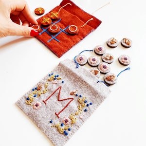 May include: A felt game set with a brown felt bag embroidered with a large "M" and floral designs. The bag contains small felt circles in various colours. A second brown felt bag is embroidered with a noughts and crosses board and contains small felt circles with star designs.