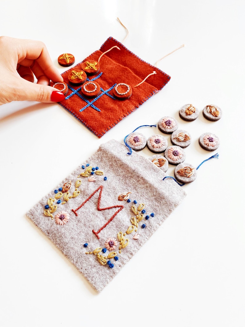 May include: A felt game set with a brown felt bag embroidered with a large "M" and floral designs. The bag contains small felt circles in various colours. A second brown felt bag is embroidered with a noughts and crosses board and contains small felt circles with star designs.