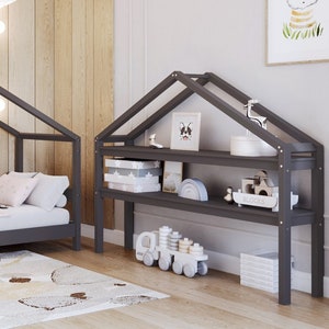 Könnte beinhalten: Ein graues Kinderbett aus Holz mit einem hausförmigen Baldachin und einer grauen Matratze. Das Bett ist mit einer braunen und weißen Decke und Kissen dekoriert. Auf der rechten Seite des Bildes befindet sich ein graues Holzregal mit zwei Regalbrettern. Das Regal ist mit Büchern, Spielzeug und einem kleinen gerahmten Bild dekoriert.