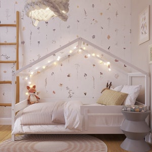 May include: White wooden house-shaped bed frame with a white mattress, white bedding, and a stuffed fox toy. The bed is decorated with fairy lights and a floral wallpaper.