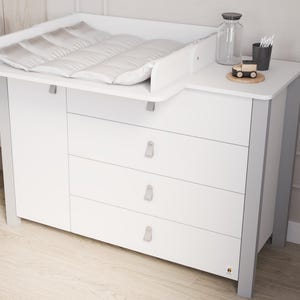 Könnte beinhalten: Ein weiß-grauer Wickeltisch mit Wickelauflage, Schubladen und einem Schrank. Der Wickeltisch hat einen grauen Rahmen und silberne Schubladengriffe. Im Hintergrund ist ein Kinderbett zu sehen.
