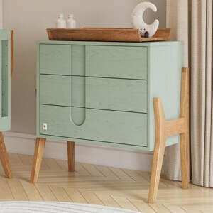 May include: A mint green chest of drawers with three drawers and wooden legs, part of a nursery set. A wooden shelf with a stuffed toy and a decorative moon are also visible. The room has a light feel with a round rug and a cot.