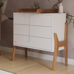 Può includere: Una moderna cameretta con una culla e un comò bianchi. Il comò ha tre cassetti e gambe in legno. Un vassoio di legno è posto sopra. La parete presenta un murale di gru e rami. Un tappeto bianco e pavimento in legno completano la scena.