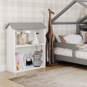 Könnte beinhalten: Ein graues, hausförmiges Bettgestell aus Holz mit einer weißen Matratze und einem weißen Bücherregal mit Büchern und einer Giraffenfigur. Die Wand hinter dem Bett hat eine Tapete mit Karomuster und ein Aquarellbild von Giraffen.