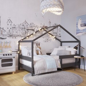May include: A gray wooden bed frame with a house-shaped canopy, featuring a white and gray patterned duvet and pillows. The bed is decorated with string lights and a dreamcatcher. The room has a white wall with a black and white illustration of a cityscape.
