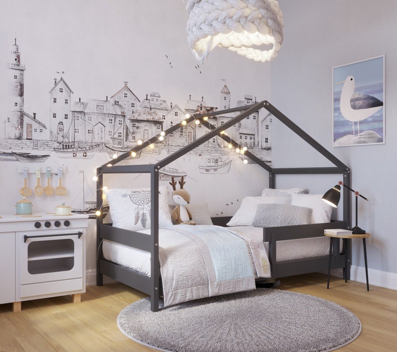 May include: A gray wooden bed frame with a house-shaped canopy, featuring a white and gray patterned duvet and pillows. The bed is decorated with string lights and a dreamcatcher. The room has a white wall with a black and white illustration of a cityscape.