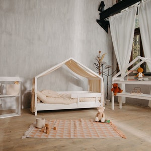 May include: A white wooden playhouse bed with a canopy and a white wooden bookshelf with shelves for toys. The bed has a white mattress and a white pillow. The bookshelf has a teddy bear, books, and other toys on the shelves. The room has a light gray wall and a wooden floor.