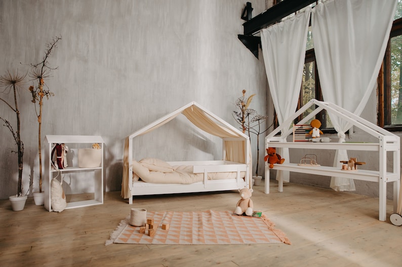May include: A white wooden playhouse bed with a canopy and a white wooden bookshelf with shelves for toys. The bed has a white mattress and a white pillow. The bookshelf has a teddy bear, books, and other toys on the shelves. The room has a light gray wall and a wooden floor.