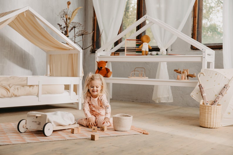 May include: A young girl plays with wooden blocks in a room with a white playhouse and a white canopy bed. The playhouse has shelves with toys, including a teddy bear, a wooden abacus, and a wooden toy.