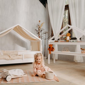 May include: A young girl plays with wooden blocks in a white playroom. The room features a white canopy bed with a white and beige bedding set, a white playhouse with shelves, and a white rug with a pink geometric pattern.
