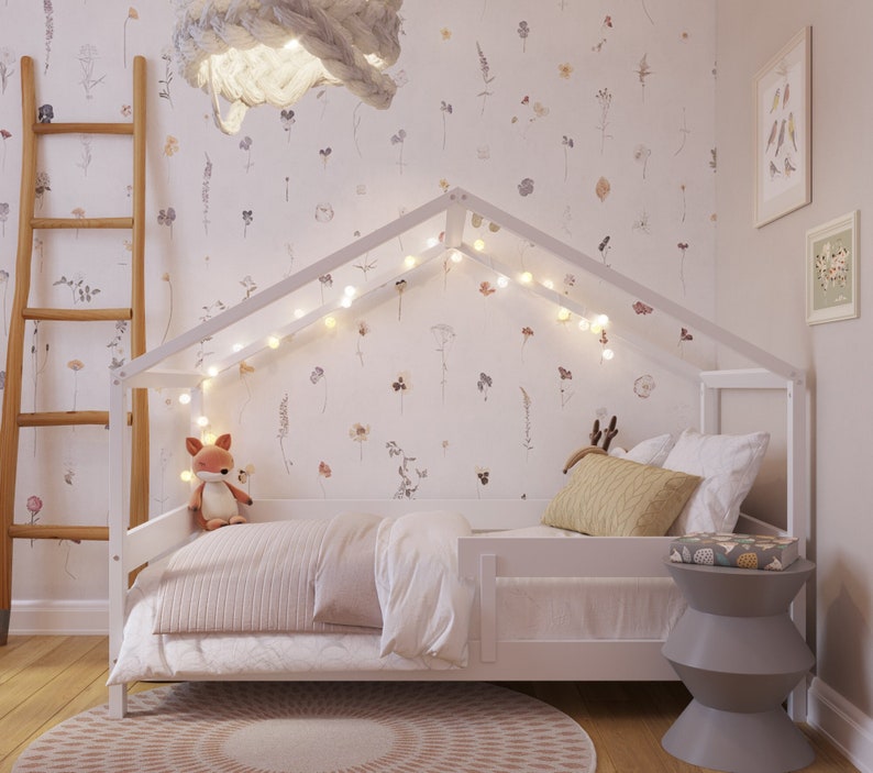 May include: White wooden house-shaped bed frame with a white mattress, white bedding, and a stuffed fox toy. The bed is decorated with fairy lights and a floral wallpaper.