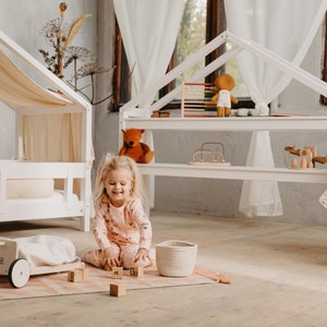 Könnte beinhalten: Ein junges Mädchen spielt mit Holzklötzen in einem Raum mit einem weißen Spielhaus und einem weißen Himmelbett. Das Spielhaus hat Regale mit Spielzeug, darunter ein Teddybär, ein Holzkugelbahn und ein Holzspielzeug.