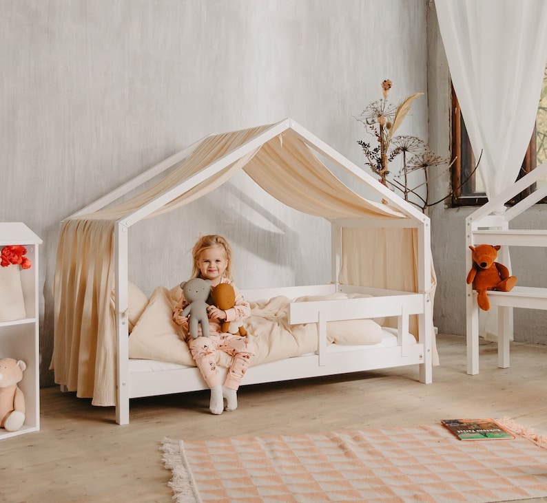May include: White wooden canopy bed with a white canopy and a girl sitting on the bed holding two stuffed animals. The bed has a white mattress and a white headboard. The bed is in a room with a white wall and a rug on the floor.