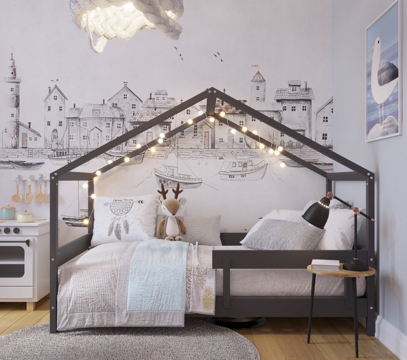 May include: A gray wooden bed frame with a house-shaped canopy and a white and gray bedding set. The bed is decorated with string lights and a stuffed deer toy. The wall behind the bed features a black and white illustration of a cityscape.