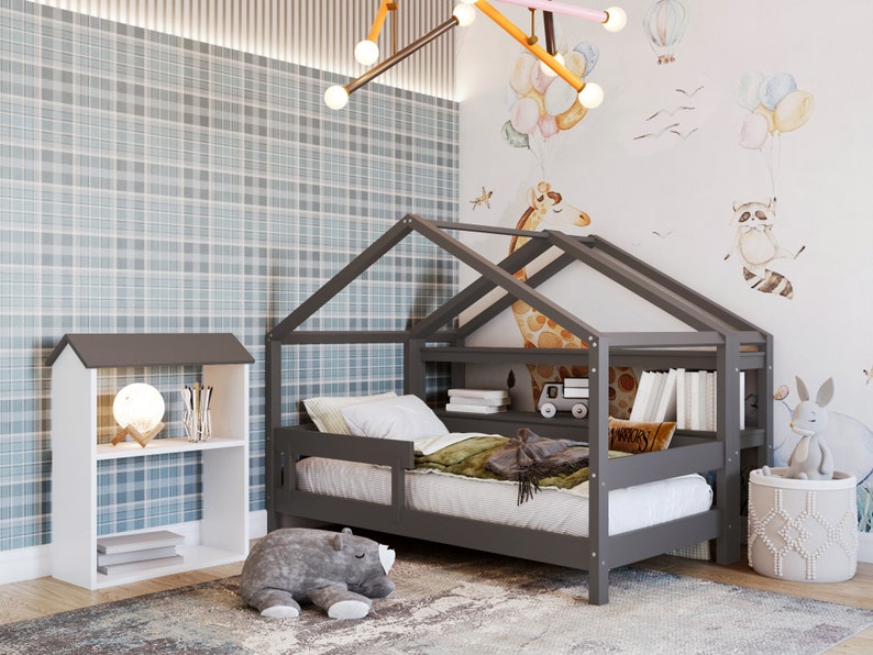 May include: A gray wooden house-shaped bed frame with a white mattress and a gray and white bookshelf next to it. The bed has a built-in shelf and a small toy truck on it. The wall behind the bed has a light blue and white plaid pattern and a white wall with a watercolor illustration of a giraffe, a raccoon, and balloons.