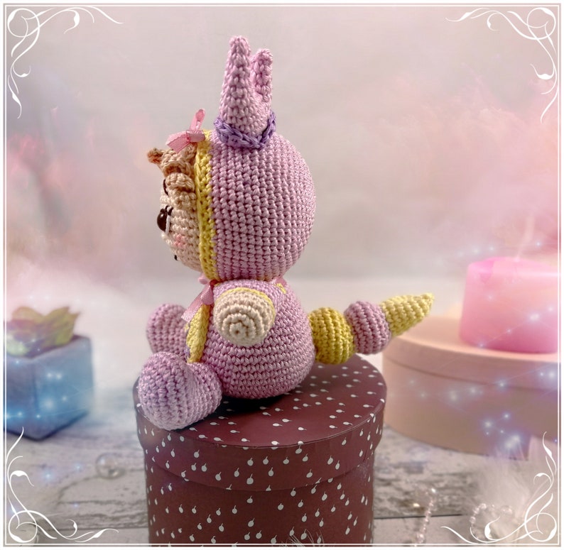 May include: A pink crocheted stuffed animal wearing a yellow and pink crown. The animal is sitting on a brown box with white polka dots.