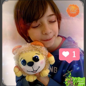 May include: A child holds a crocheted lion plush toy. The lion is yellow and orange with black eyes and a pink nose. The child is wearing a blue shirt with a white heart icon that says "1".