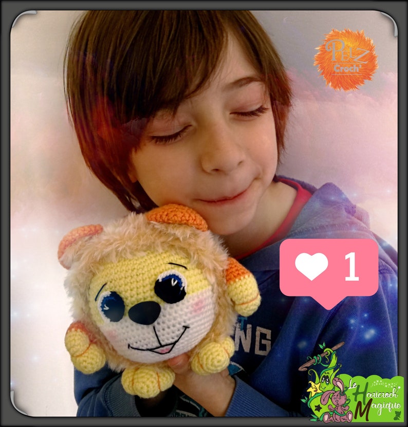 May include: A child holds a crocheted lion plush toy. The lion is yellow and orange with black eyes and a pink nose. The child is wearing a blue shirt with a white heart icon that says "1".