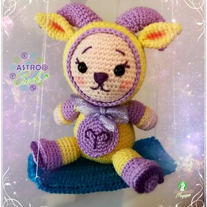May include: A crocheted amigurumi toy in the shape of an Aries zodiac sign. The toy is yellow and purple with a silver bow and is sitting on a blue and purple crocheted cushion. The toy has a cute face with black eyes and a pink nose.