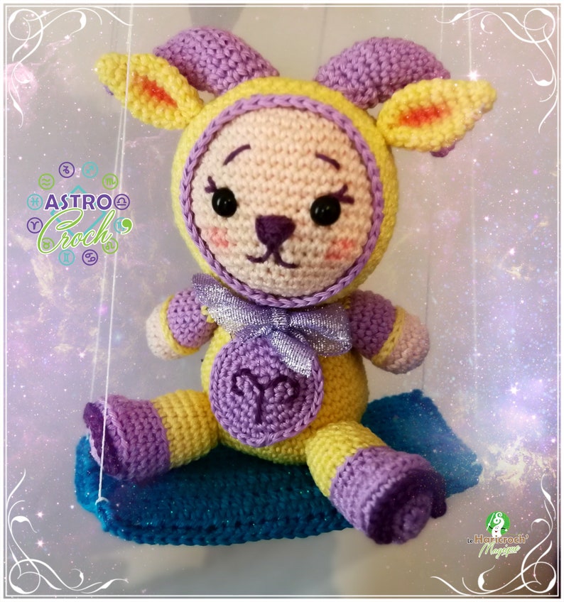 May include: A crocheted amigurumi toy in the shape of an Aries zodiac sign. The toy is yellow and purple with a silver bow and is sitting on a blue and purple crocheted cushion. The toy has a cute face with black eyes and a pink nose.