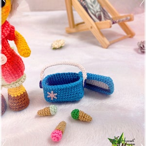 May include: A crocheted doll wearing a red and yellow outfit and brown boots, holding a blue crocheted cooler with a snowflake design. There are three crocheted ice cream cones on the white surface. The cooler is open and the doll is looking at the ice cream cones.