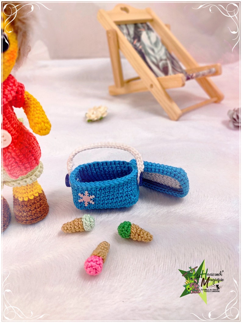 May include: A crocheted doll wearing a red and yellow outfit and brown boots, holding a blue crocheted cooler with a snowflake design. There are three crocheted ice cream cones on the white surface. The cooler is open and the doll is looking at the ice cream cones.